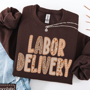 May include: A dark chocolate brown crewneck sweatshirt featuring the text LABOR DELIVERY in large, blocky letters. The letters have a mixed pattern design including leopard print, beige gingham, and a floral motif. A silver chain necklace rests on top.
