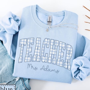 May include: A pale blue sweatshirt featuring the word "TEACHER" in a blue and white gingham design. The name "Mrs. Adams" is printed below. A silver necklace is draped on the sweatshirt.