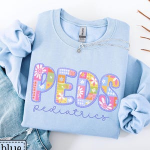 May include: Light blue sweatshirt with the word "PEDS" in colorful floral letters. The word "pediatrics" is written below in cursive. The sweatshirt is folded on top of a pair of blue jeans.