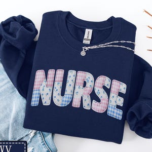 May include: A navy blue sweatshirt featuring the word "NURSE" in a patchwork design of pink, blue, and white floral and gingham patterns. A silver necklace with a pendant rests on the sweatshirt. The word "navy" is in a navy blue rectangle.