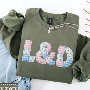 May include: A military green sweatshirt with the letters "L & D" appliqued in a patchwork design of pink and blue floral and gingham patterns. A silver necklace with a small pendant rests on the sweatshirt. Light blue denim jeans are visible in the background.