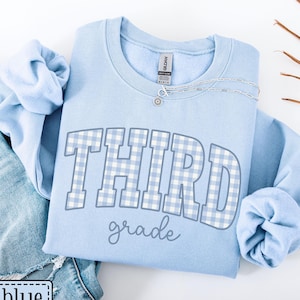 May include: Light blue sweatshirt with the text "THIRD grade" in a white and light blue gingham pattern. The sweatshirt is folded on top of a pair of blue jeans. A silver necklace is draped across the sweatshirt. The words "light blue" are in a white box.