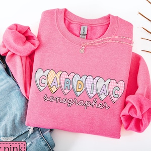 Peut inclure: Un sweat-shirt rose sécurité avec l'inscription "CARDIAC sonographer" dans un motif en forme de cœur. Le sweat-shirt est associé à un jean en denim bleu. Un collier délicat est drapé sur le sweat-shirt.