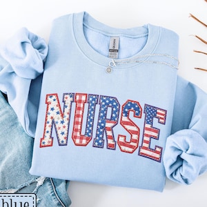 May include: A light blue sweatshirt with the word "NURSE" in a patriotic design featuring red, white, and blue stars and stripes. The sweatshirt is paired with blue jeans. A silver necklace is draped on the sweatshirt. The text "light blue" is in a white box.