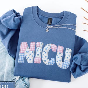 May include: A blue crew neck sweatshirt with the word "NICU" appliqued on the front in a patchwork design. The letters feature floral, gingham, and pink patterns. A silver necklace rests on the shirt. The word "indigo" is in a blue rectangle in the bottom left corner.