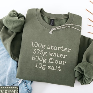 May include: An olive green sweatshirt with white text detailing baking ingredients: 100g starter, 375g water, 500g flour, and 10g salt. A silver necklace is on the shirt. Light blue denim jeans are also visible.