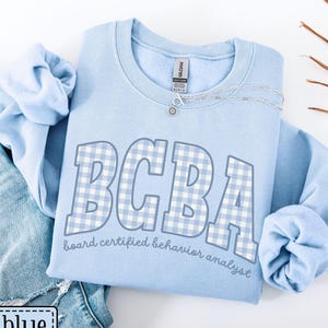 May include: A light blue sweatshirt with the letters "BCBA" in a light blue and white gingham pattern. The words "board certified behavior analyst" are printed below the letters. The sweatshirt is paired with blue jeans.