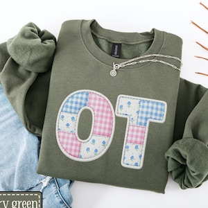 May include: Military green sweatshirt with the letters "OT" appliquéd on the front. The letters are made of patchwork fabric in pink, blue, and white floral and gingham patterns. A silver necklace is draped on the sweatshirt, which is paired with blue jeans.