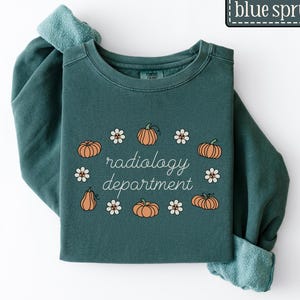 May include: A teal sweatshirt featuring the words "radiology department" in a script font, with pumpkin and daisy illustrations. The text "blue spruce" is in a rectangular box at the top right of the image.