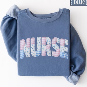 May include: A blue jean colored sweatshirt with the word "NURSE" in patchwork letters. The letters are a mix of pink gingham, blue floral, and blue gingham patterns. The sweatshirt has long sleeves.