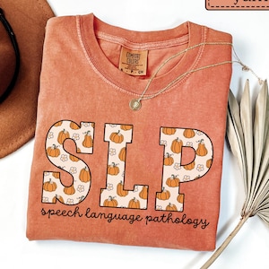 May include: A coral-colored t-shirt with the letters "SLP" in a pumpkin pattern. The words "speech language pathology" are printed below the letters. A gold necklace and a brown hat are also visible.
