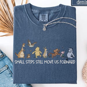 May include: A denim-colored short-sleeve t-shirt featuring a graphic of Winnie the Pooh characters walking in a line. Below the characters, the text reads, "SMALL STEPS STILL MOVE US FORWARD." The shirt has a crew neckline and a Comfort Colors brand label.