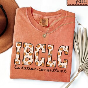 May include: A coral-colored t-shirt with the text "IBCLC lactation consultant" in a pumpkin-themed design. The shirt has a round neck and short sleeves. A gold necklace and a brown hat are also in the image.