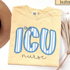May include: A butter yellow short-sleeve t-shirt featuring the text ICU in large, light blue and white striped letters with a scalloped edge and a small blue bow graphic on the letter C. The word nurse is printed below in black cursive script.