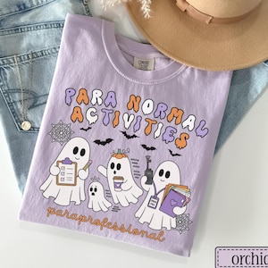May include: An orchid-colored t-shirt featuring a graphic with three white ghosts and the text "PARA NORMAL ACTIVITIES" and "paraprofessional". The ghosts are depicted with a clipboard, a coffee cup, a walkie-talkie, and a stack of folders.