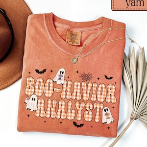May include: A yam-colored t-shirt featuring the text BOO-HAVIOR ANALYST in a white and orange gingham pattern. The design includes three white ghosts, black bats, and a spider web. The shirt has a crew neckline and a Comfort Colors brand label.