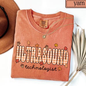 Puede incluir: Una camiseta color &ntilde;ame con las palabras "ULTRASOUND technologist" en un dise&ntilde;o a cuadros. La camiseta presenta gr&aacute;ficos con tem&aacute;tica oto&ntilde;al de bellotas y hojas. Tambi&eacute;n se ven un collar dorado y un sombrero marr&oacute;n.