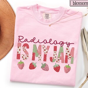 May include: A light pink t-shirt with the phrase "Radiology IS MY JAM" in a patchwork design with strawberries. The shirt has a crew neck and a small tag on the upper chest.