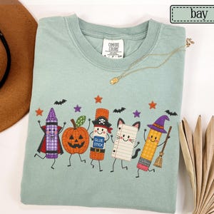 May include: A sage green crewneck t-shirt featuring a graphic print of five cartoon characters: a purple crayon, a jack-o'-lantern, a glue stick wearing a pirate hat, a piece of lined paper with cat ears, and a pencil wearing a witch hat.
