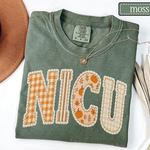 May include: A moss green t-shirt with the letters "NICU" appliqued on the front. The letters are made of various autumn-themed fabrics, including orange gingham and pumpkin patterns. A gold necklace rests on the shirt.