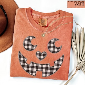 May include: A short-sleeved crew neck t-shirt in a solid yam orange color. The front features a large jack-o'-lantern face applique made of black and white buffalo plaid fabric with a stitched border. The shirt has a Comfort Colors brand label.