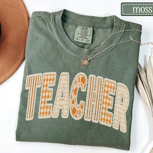May include: A moss green Comfort Colors t-shirt with the word "TEACHER" appliqued across the front in a patchwork design. The letters feature various patterns, including stripes, gingham, and floral prints. A gold necklace rests on the shirt.