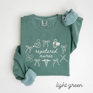 May include: A light green sweatshirt with the words "registered nurse" in white script, surrounded by medical-themed illustrations. The sweatshirt has a crew neck and long sleeves with a light blue lining. The words "light green" are written in script at the bottom.