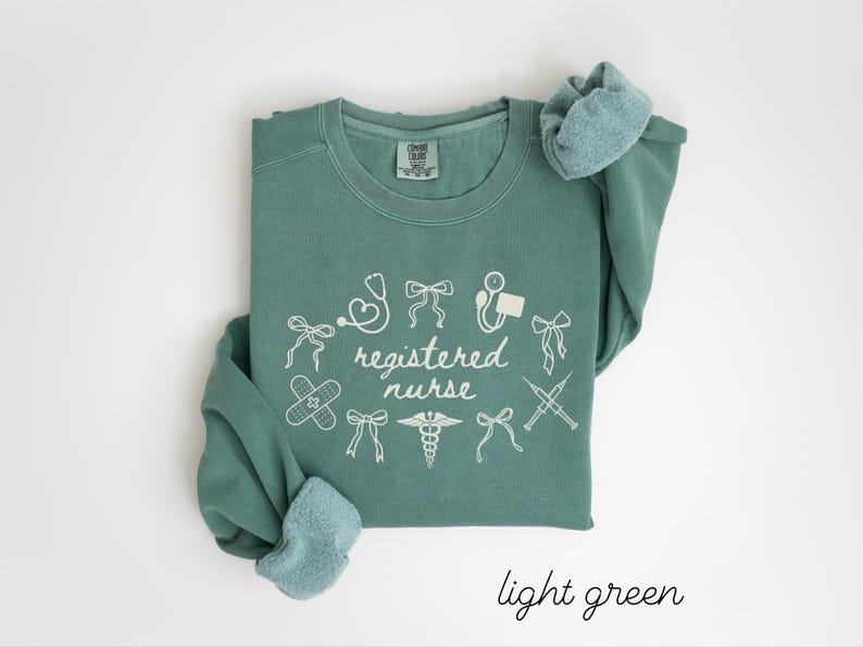 May include: A light green sweatshirt with the words "registered nurse" in white script, surrounded by medical-themed illustrations. The sweatshirt has a crew neck and long sleeves with a light blue lining. The words "light green" are written in script at the bottom.