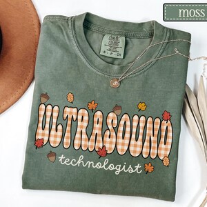 Puede incluir: Una camiseta verde musgo con las palabras "ULTRASOUND technologist" en una fuente retro con un patr&oacute;n de cuadros. La camiseta est&aacute; decorada con hojas de oto&ntilde;o y bellotas. Un collar plateado con un colgante est&aacute; drapeado sobre la camiseta.
