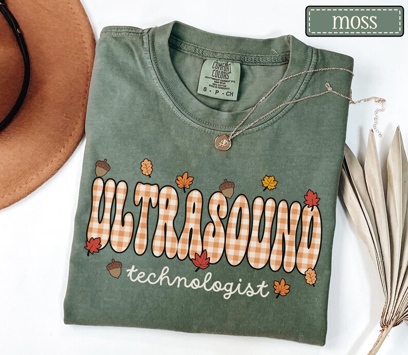 Puede incluir: Una camiseta verde musgo con las palabras "ULTRASOUND technologist" en una fuente retro con un patr&oacute;n de cuadros. La camiseta est&aacute; decorada con hojas de oto&ntilde;o y bellotas. Un collar plateado con un colgante est&aacute; drapeado sobre la camiseta.