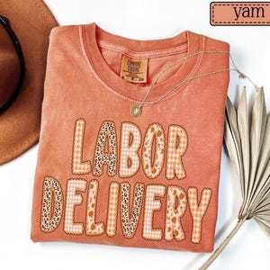 May include: A yam-coloured short-sleeve t-shirt with a crew neckline. The front features the text LABOR DELIVERY in large, blocky letters filled with a mix of leopard print, orange gingham, and autumn-themed patterns including small pumpkins and leaves.