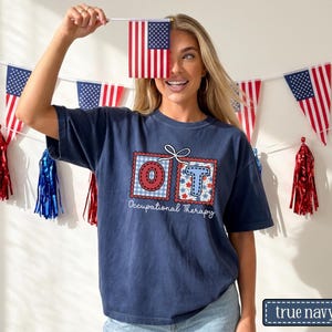 May include: Navy blue t-shirt with a graphic that says "OT Occupational Therapy" in a red and blue design. The image also includes American flag decorations and a small American flag.
