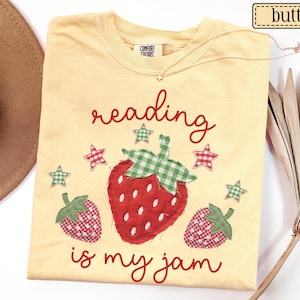 May include: A butter yellow t-shirt featuring a red strawberry graphic with a green gingham top, two smaller strawberries, and five gingham stars. The text reads reading is my jam in red cursive script. The shirt has a Comfort Colors brand label.