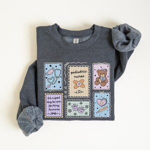 May include: A dark gray sweatshirt with a colorful graphic design. The design includes illustrations of a stethoscope, band-aid, teddy bear, and a heart with a thermometer. Text on the sweatshirt reads "pediatric nurse" and "It's a good day to care for tiny humans."