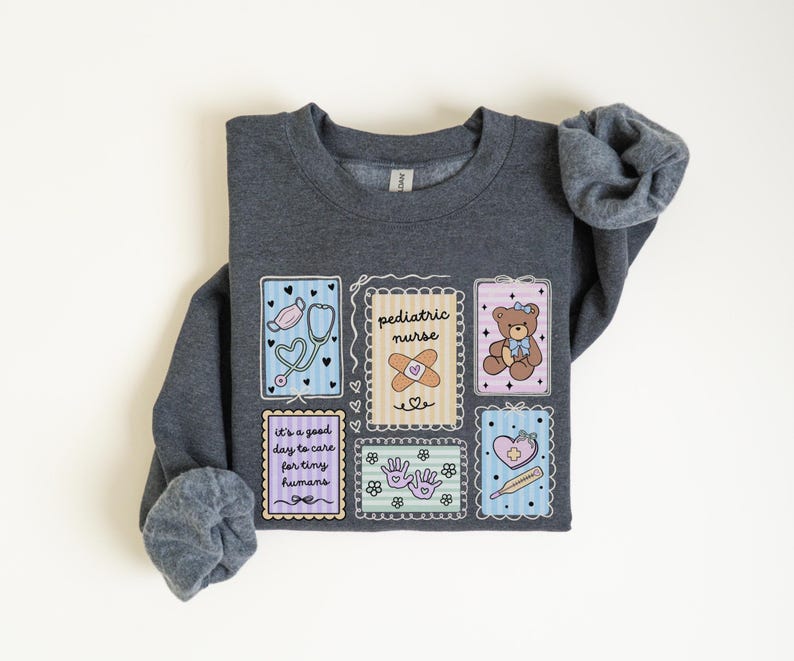 May include: A dark gray sweatshirt with a colorful graphic design. The design includes illustrations of a stethoscope, band-aid, teddy bear, and a heart with a thermometer. Text on the sweatshirt reads "pediatric nurse" and "It's a good day to care for tiny humans."