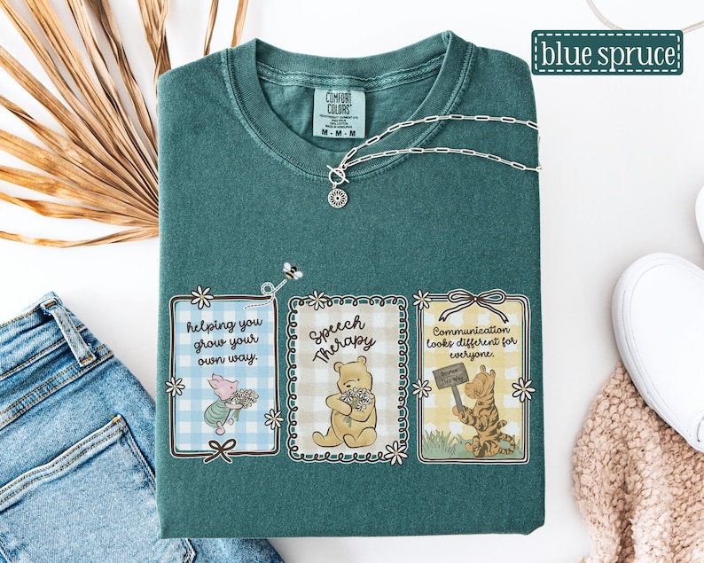May include: A teal Comfort Colors t-shirt with three framed illustrations featuring Winnie the Pooh characters. The illustrations include text that reads "helping you grow your own way," "Speech Therapy," and "Communication looks different for everyone." A silver necklace is draped across the shirt.