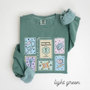 Pode incluir: Camisola verde claro com patches de tem&aacute;tica m&eacute;dica. Os patches incluem um estetosc&oacute;pio, um s&iacute;mbolo m&eacute;dico, um kit de primeiros socorros, uma ligadura e um term&oacute;metro. O texto "emergency department" tamb&eacute;m est&aacute; presente. As palavras "light green" est&atilde;o escritas em it&aacute;lico na parte inferior.