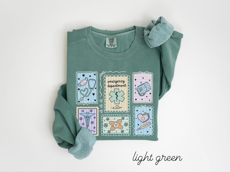 Pode incluir: Camisola verde claro com patches de tem&aacute;tica m&eacute;dica. Os patches incluem um estetosc&oacute;pio, um s&iacute;mbolo m&eacute;dico, um kit de primeiros socorros, uma ligadura e um term&oacute;metro. O texto "emergency department" tamb&eacute;m est&aacute; presente. As palavras "light green" est&atilde;o escritas em it&aacute;lico na parte inferior.