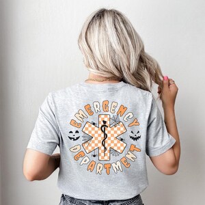 Op de afbeelding: Een lichtgrijs t-shirt met korte mouwen en een afbeelding op de achterkant. Het ontwerp bevat een oranje en wit geblokt Ster van het Leven-symbool met een zwarte esculaap, omringd door de tekst EMERGENCY DEPARTMENT en spinnenweb-iconen.
