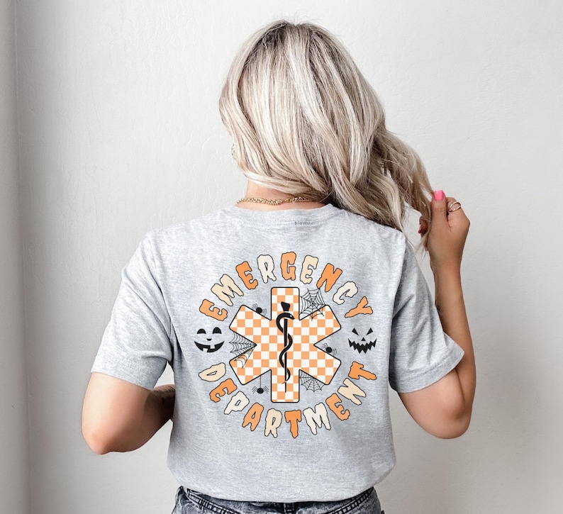 Op de afbeelding: Een lichtgrijs t-shirt met korte mouwen en een afbeelding op de achterkant. Het ontwerp bevat een oranje en wit geblokt Ster van het Leven-symbool met een zwarte esculaap, omringd door de tekst EMERGENCY DEPARTMENT en spinnenweb-iconen.
