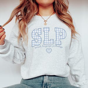 May include: A light gray sweatshirt with a blue and white gingham pattern spelling "SLP" and the text "speech language pathology" below. A small blue heart is centered below the text.