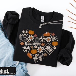May include: A black long-sleeve sweatshirt featuring a heart-shaped graphic composed of pumpkins, daisies, autumn leaves, radiation symbols, a clipboard, a skeleton hand, a medical caduceus, and an X-ray image with the text Radiology in the center.