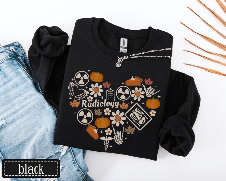 May include: A black long-sleeve sweatshirt featuring a heart-shaped graphic composed of pumpkins, daisies, autumn leaves, radiation symbols, a clipboard, a skeleton hand, a medical caduceus, and an X-ray image with the text Radiology in the center.