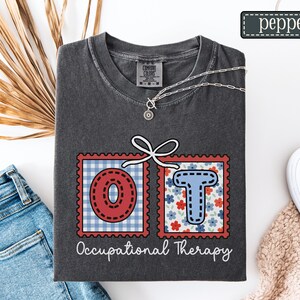 May include: Dark gray t-shirt with the letters "OT" in red and blue postage stamp designs, connected by a white bow. The words "Occupational Therapy" are written below the letters. A silver necklace and the word "pepper" are also visible.