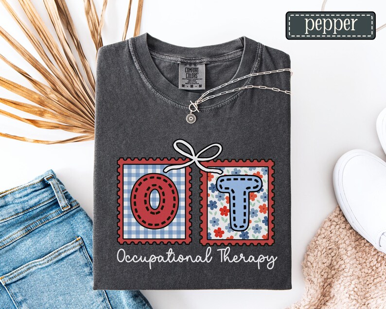 May include: Dark gray t-shirt with the letters "OT" in red and blue postage stamp designs, connected by a white bow. The words "Occupational Therapy" are written below the letters. A silver necklace and the word "pepper" are also visible.