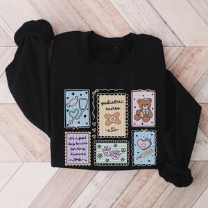 May include: Black sweatshirt with colorful patches featuring medical and care-themed illustrations. Patches include a stethoscope, band-aid, teddy bear, and text that reads "pediatric nurse" and "It's a good day to care for tiny humans."