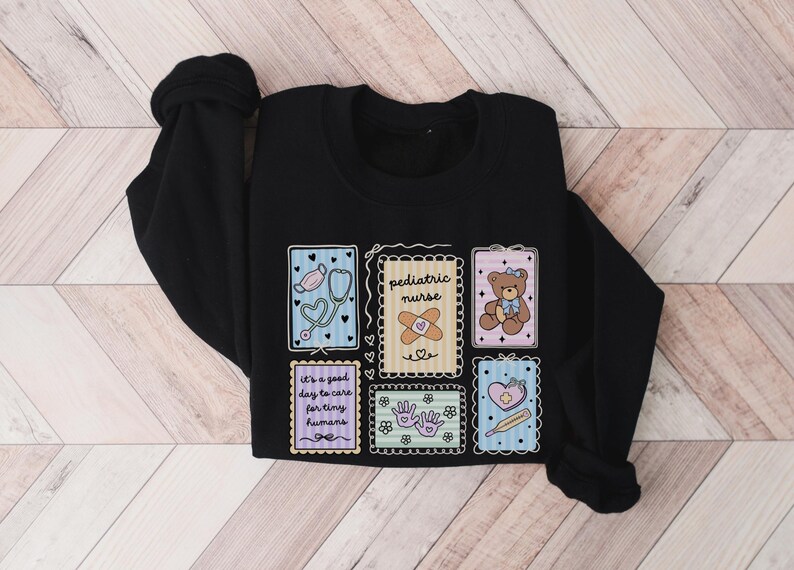 May include: Black sweatshirt with colorful patches featuring medical and care-themed illustrations. Patches include a stethoscope, band-aid, teddy bear, and text that reads "pediatric nurse" and "It's a good day to care for tiny humans."