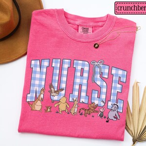 K&ouml;nnte beinhalten: Ein leuchtend pinkfarbenes T-Shirt mit dem Wort "NURSE" in blau-wei&szlig; karierten Buchstaben. Das Shirt zeigt Zeichentrickfiguren und eine goldene Halskette. Der Markenname "crunchberry" befindet sich oben rechts.