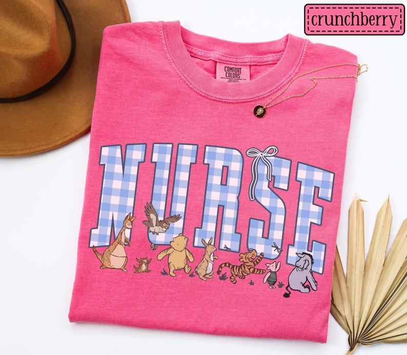 K&ouml;nnte beinhalten: Ein leuchtend pinkfarbenes T-Shirt mit dem Wort "NURSE" in blau-wei&szlig; karierten Buchstaben. Das Shirt zeigt Zeichentrickfiguren und eine goldene Halskette. Der Markenname "crunchberry" befindet sich oben rechts.