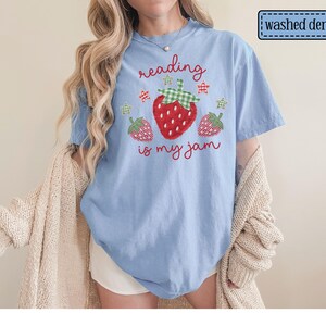 May include: A light blue washed denim t-shirt featuring a graphic of three strawberries with green gingham leaves and small stars. The text "reading is my jam" is printed in red cursive script above and below the central strawberry illustration.